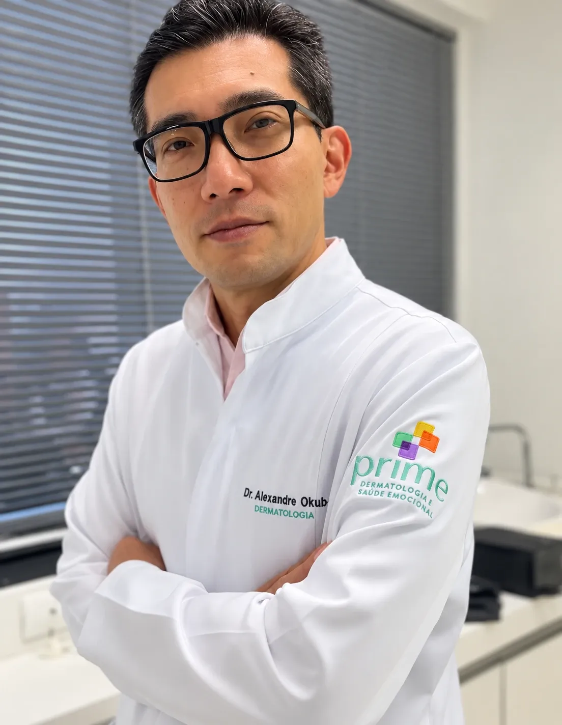 Dr. Alexandre Okubo, dermatologista, de braços cruzados e jaleco da Prime Dermatologia no consultório